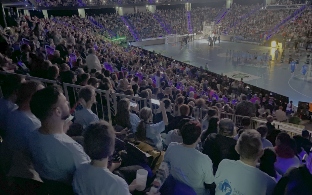 Eine große Menschenmenge füllt die Sitze in einer Sporthalle und verfolgt ein Spiel auf einem hell erleuchteten Platz. Die Zuschauer im Vordergrund tragen weiße Hemden, während die Tribünen und die Arena mit lila und blauen Lichtern beleuchtet sind.