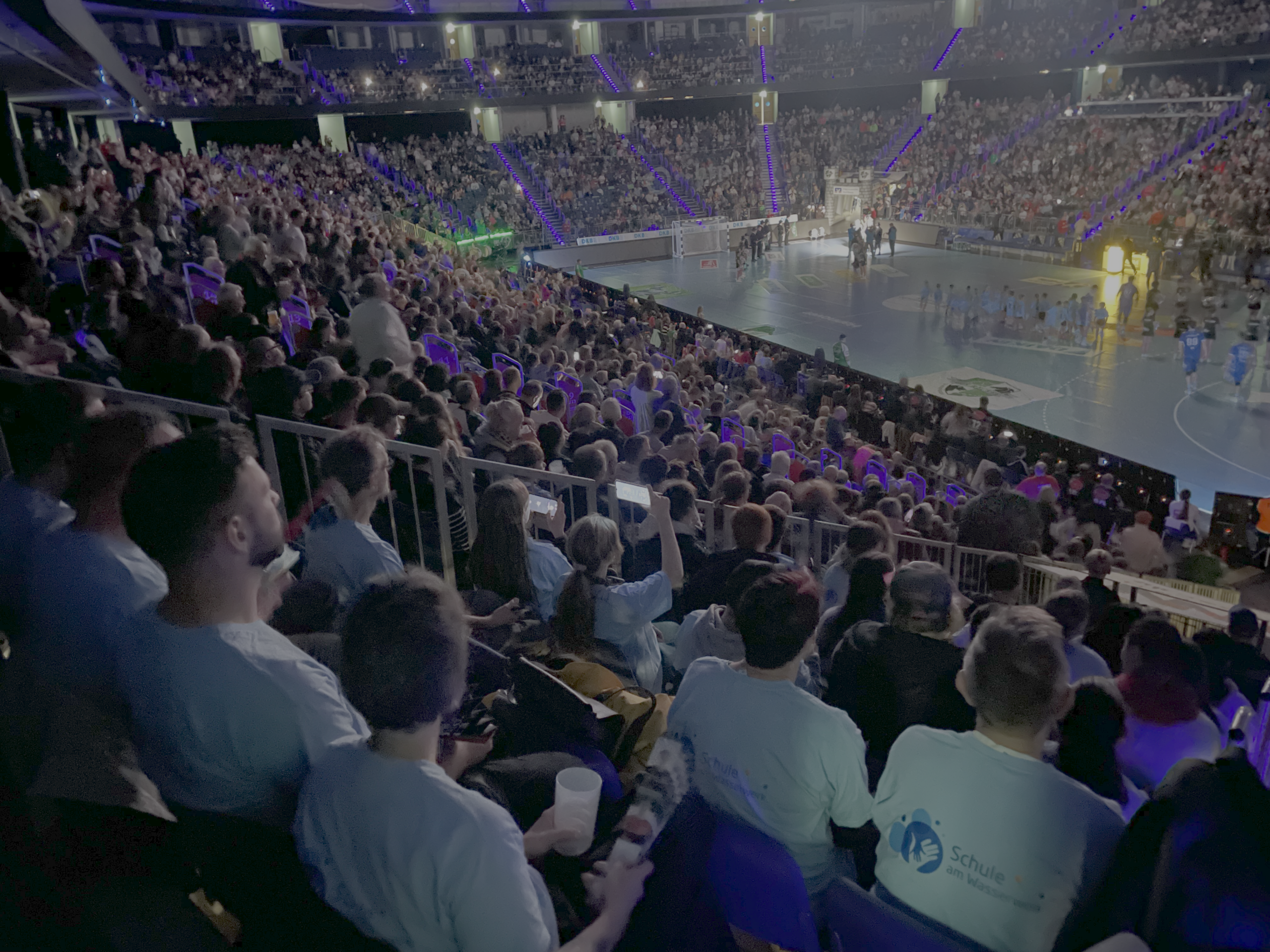 Eine große Menschenmenge füllt die Sitze in einer Sporthalle und verfolgt ein Spiel auf einem hell erleuchteten Platz. Die Zuschauer im Vordergrund tragen weiße Hemden, während die Tribünen und die Arena mit lila und blauen Lichtern beleuchtet sind.