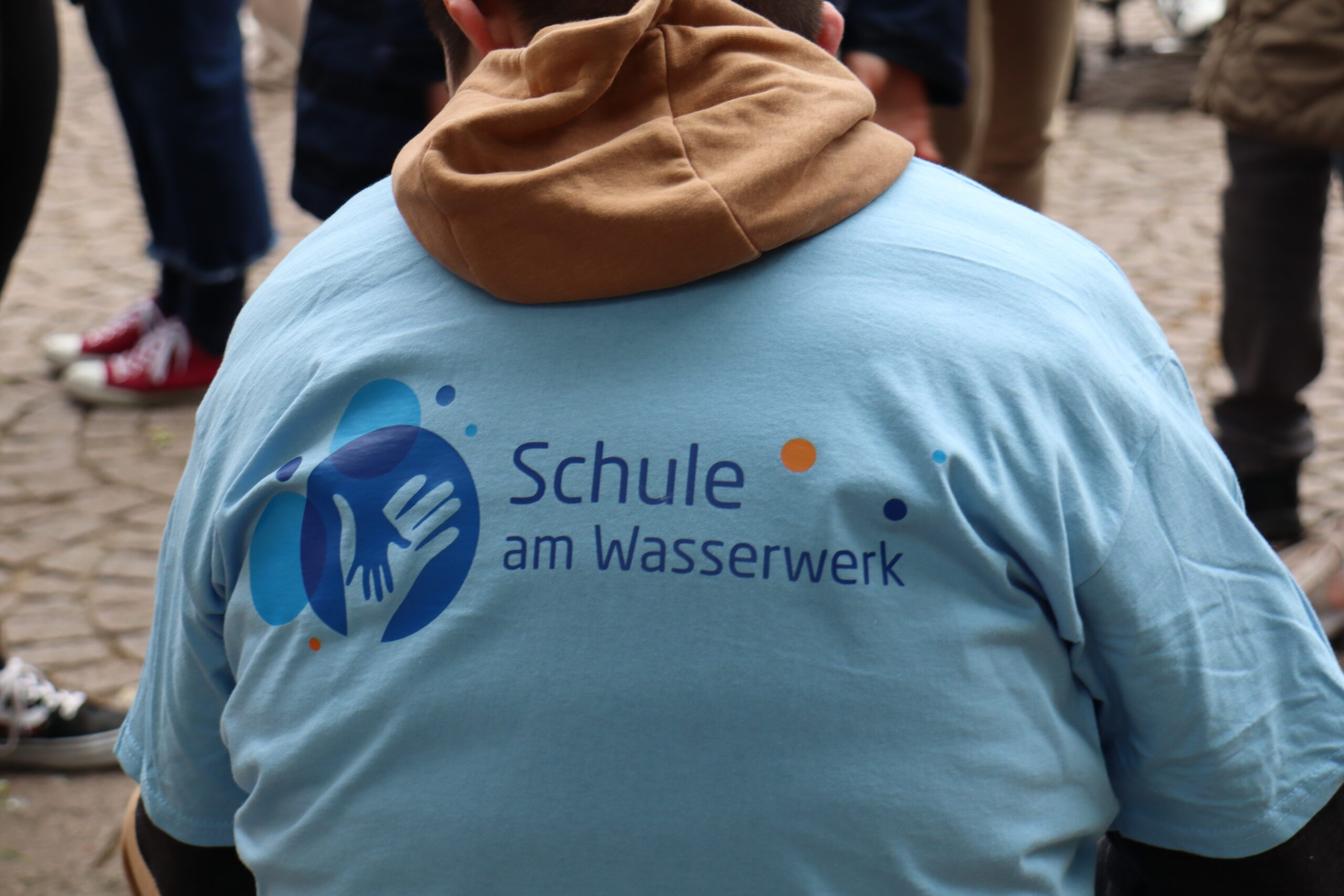 Eine Person trägt ein hellblaues Hemd mit dem Logo und Text Schule am Wasserwerk auf dem Rücken, auf dem stilisierte Wassertropfen und Hände abgebildet sind. Im Hintergrund sind weitere Personen zu sehen.